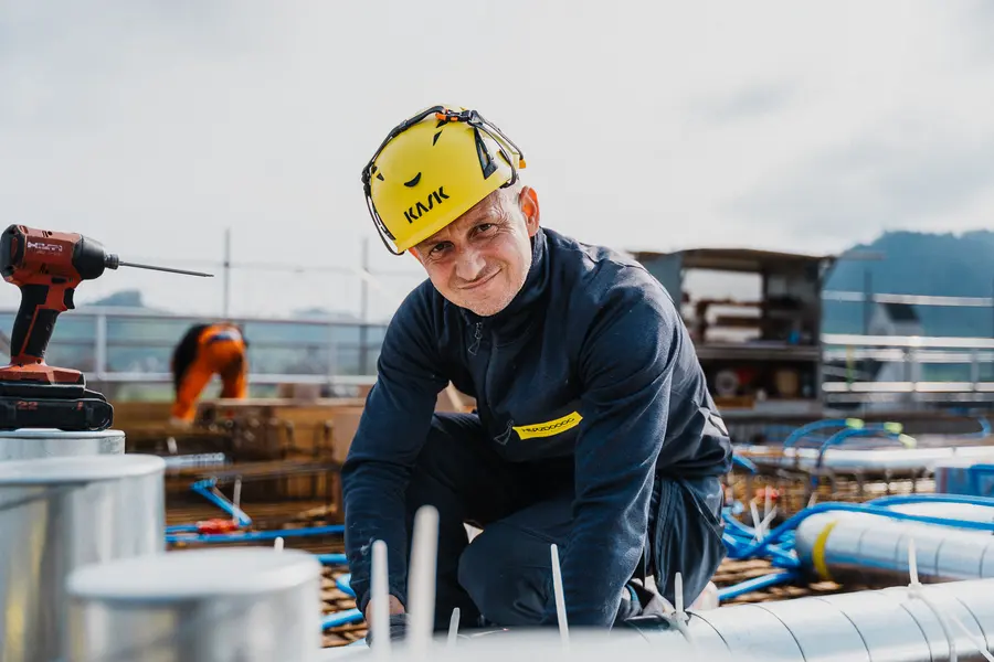 Monteur mit Helm kniet auf der Baustelle zwischen Lüftungsrohren