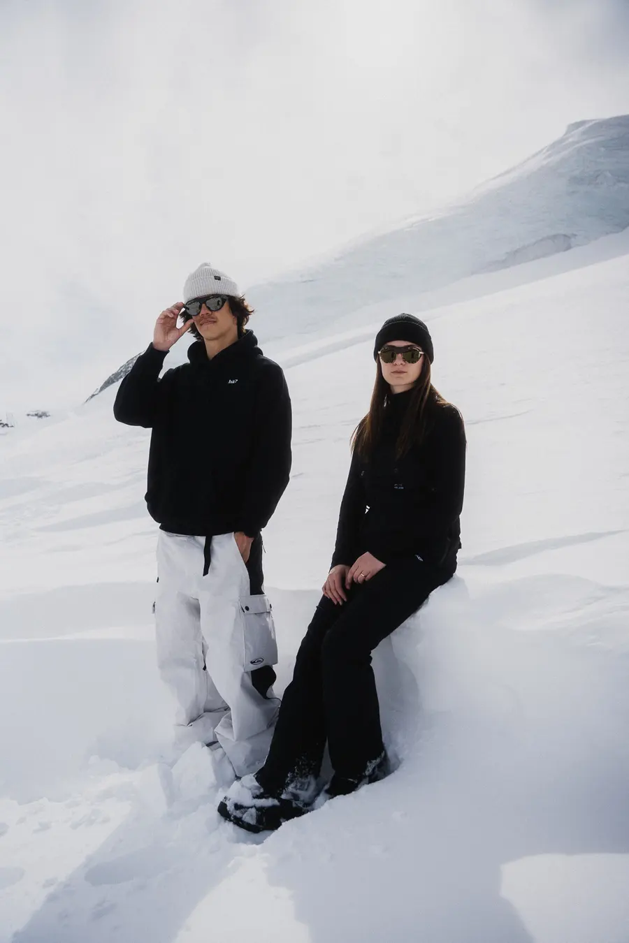 Paar in Schwarz steht und sitzt mit Sonnenbrillen im Tiefschnee