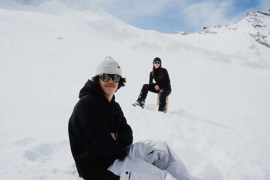 Junger Mann sitzt im Schnee, hinter ihm die Frau auf einem Pfosten