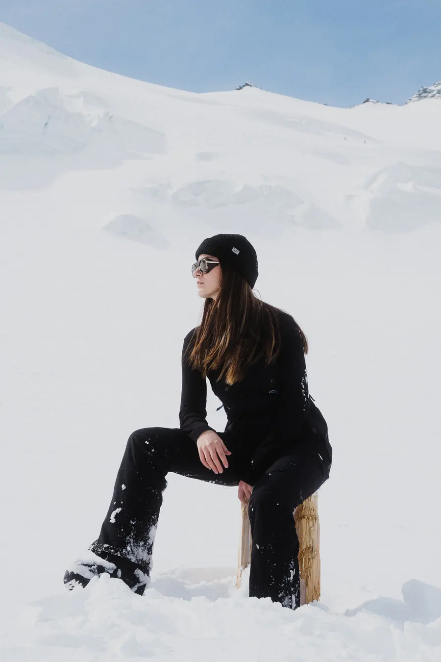 Frau in Schwarz sitzt auf einem Holzpfosten und blickt über den Schnee