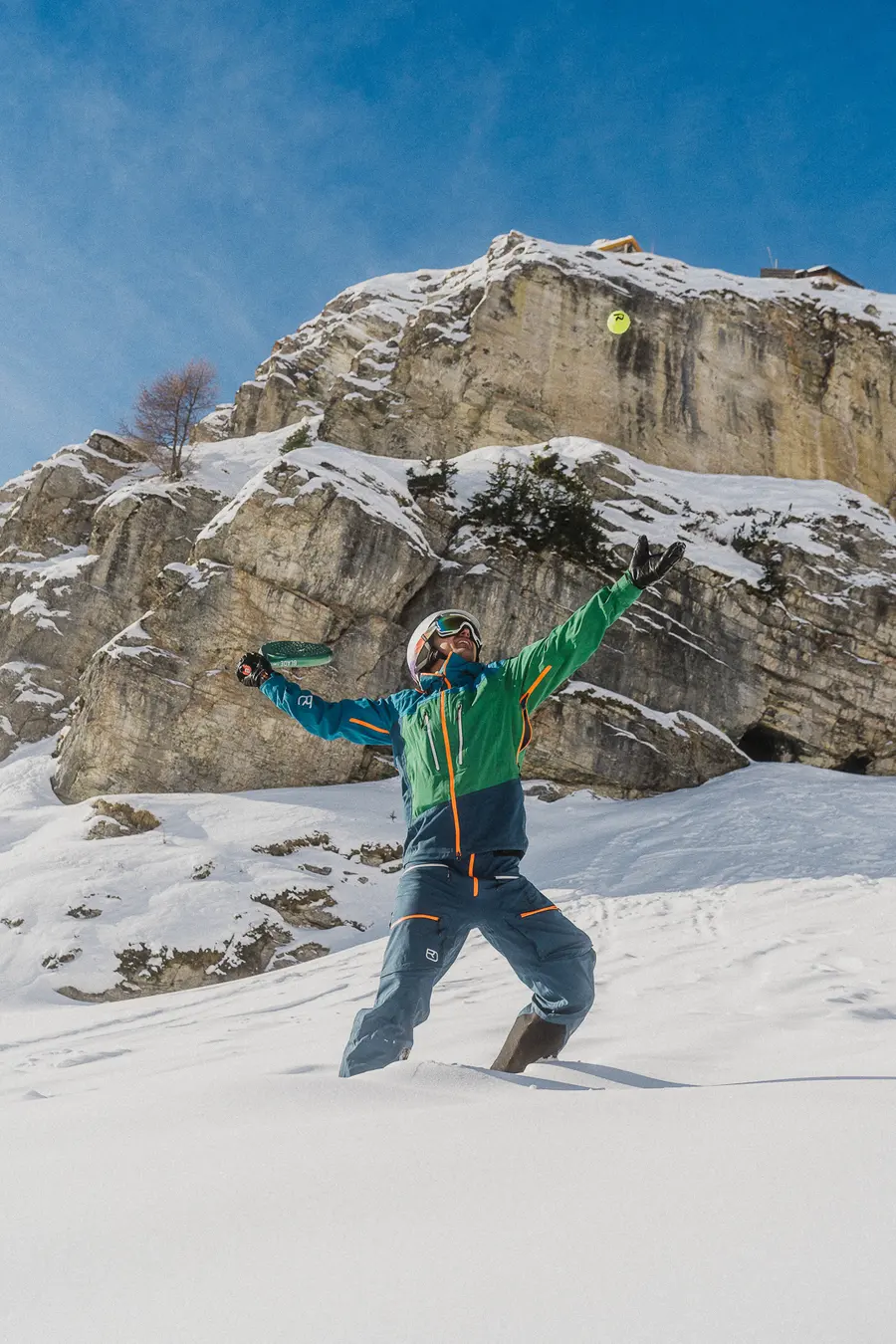 Skifahrer im grün-blauen Anzug springt vor einer verschneiten Felswand