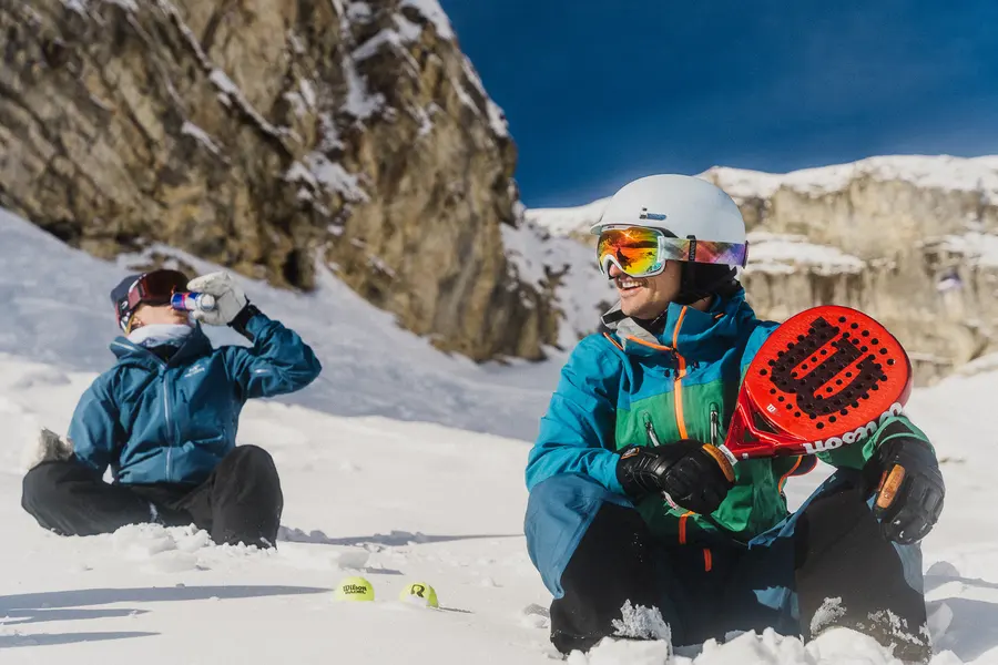 Zwei Skifahrende sitzen im Schnee, einer hält einen Padelschläger