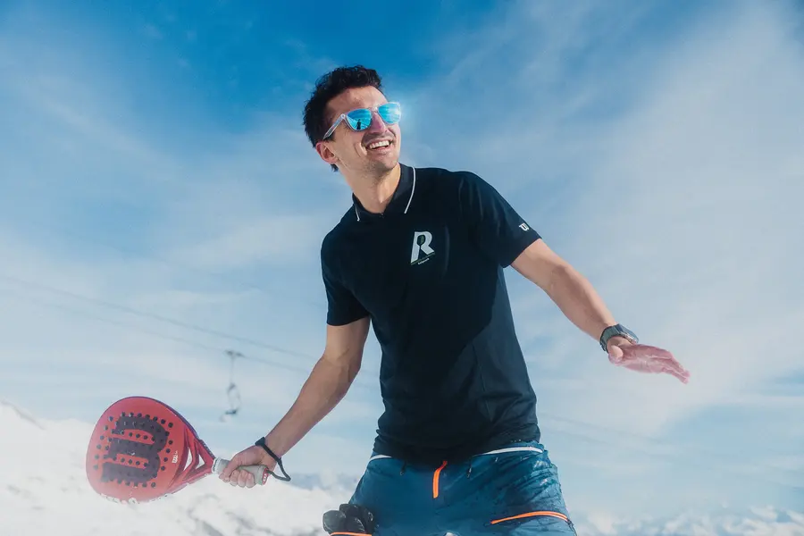 Mann im Polo mit Sonnenbrille schwingt im Schnee den Padelschläger
