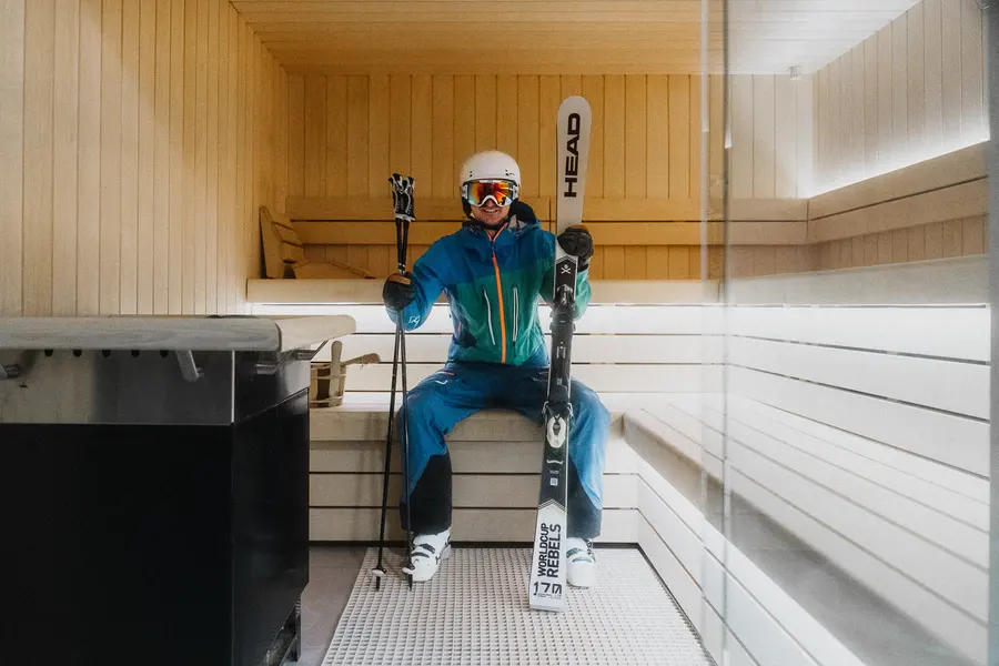 Skifahrer mit Ski und Stöcken sitzt in einer hellen Holzsauna