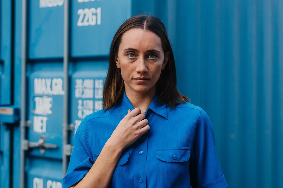 Porträt im blauen Hemd, eine Hand am Kragen, vor dem Container