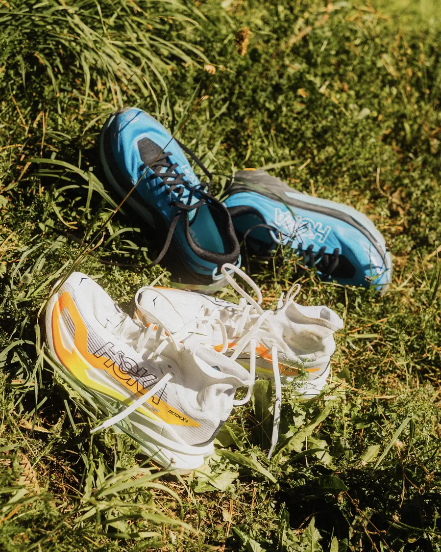 Weisse und blaue HOKA-Trailschuhe liegen im sonnigen Berggras