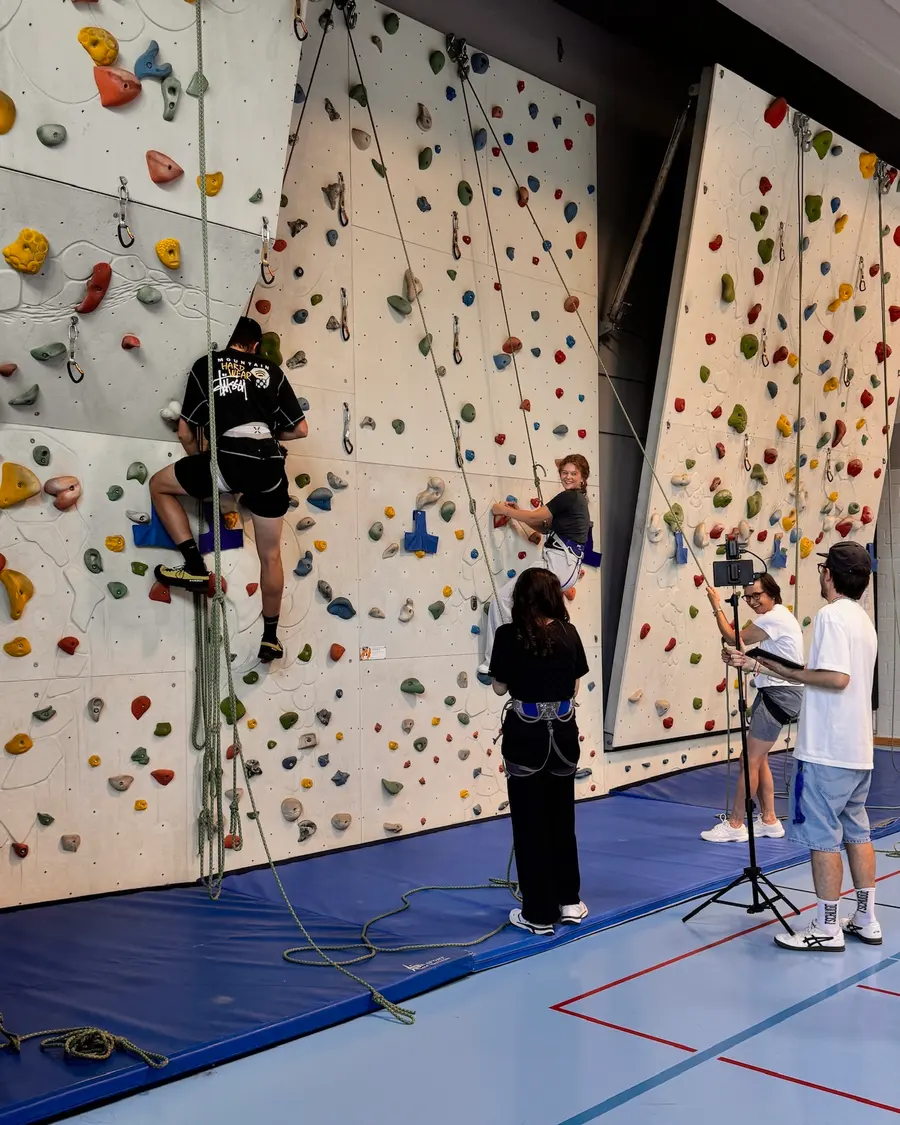 Dreh an einer Kletterwand in einer Turnhalle: Kamera auf dem Stativ, Kletternde an der Wand, das Team schaut von der Matte aus zu