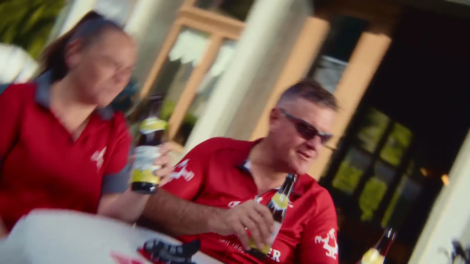Zwei Velofahrer in roten Trikots trinken Panasch vor einem Restaurant