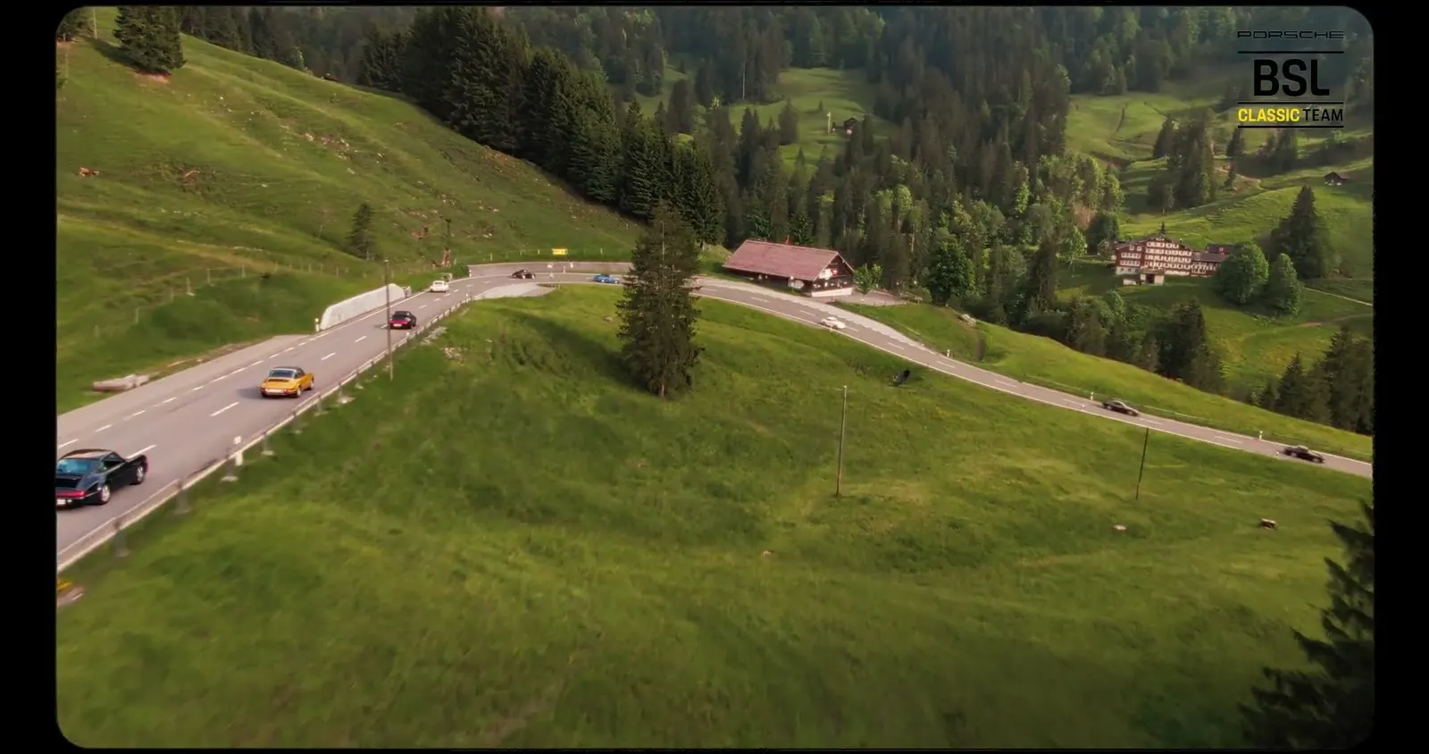 Vue aérienne d’une route de montagne avec des Porsche entre prairies et forêt