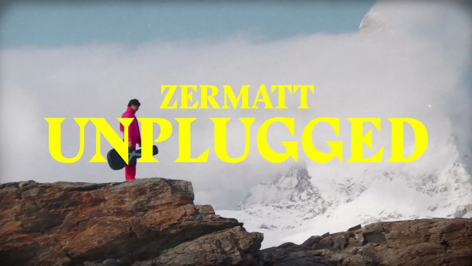 Person in Pink mit Gitarrenkoffer auf einem Felsen über dem Nebelmeer, Schriftzug Zermatt Unplugged